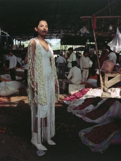 A full-length look from the 'Phool Mandi' editorial. The styling features a white fringed dress layered with a jacket made of jasmine flowers, blending fashion design with natural elements in a raw, market environment.