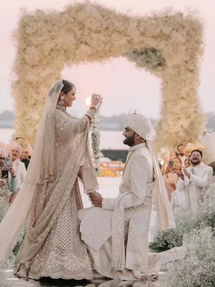 The bride places the varmala on her kneeling groom, a moment of playful tradition and deep affection. The sunset over the Kochi backwaters provides a stunning, natural backdrop to their all-white floral mandap.