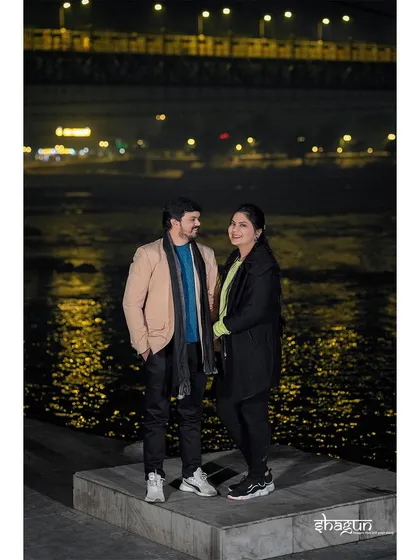 A casual, relaxed portrait by the river at night. The couple is dressed in comfortable winter wear, showing a more realistic and relatable side of their love story.