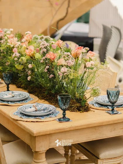 A rustic wooden table set with a lush runner of moss and wildflowers. The blue glassware and patterned plates add a touch of Mediterranean charm.