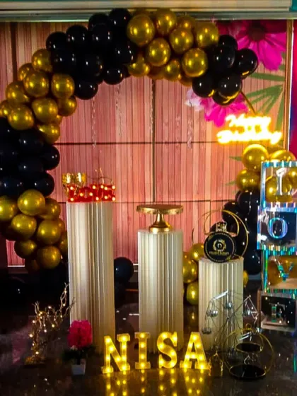 A bold and glamorous birthday setup featuring a black and metallic gold balloon arch. The decor is completed with a neon sign, illuminated name letters, and 'LOVE' blocks, all set against a warm wood-paneled wall.