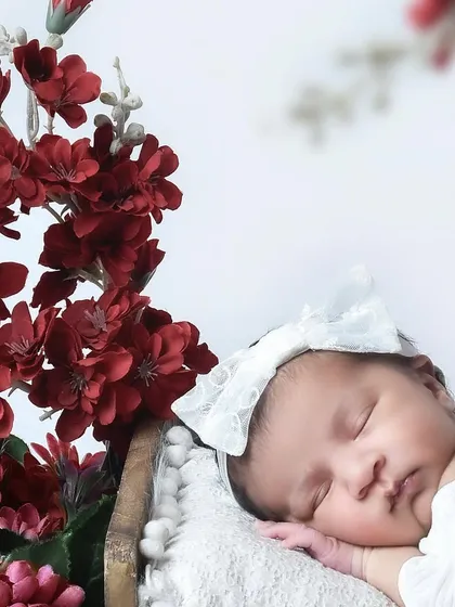 A sleeping newborn girl wearing a large white bow, posed next to a beautiful arrangement of deep red flowers. The contrast between the white and red creates a striking image.