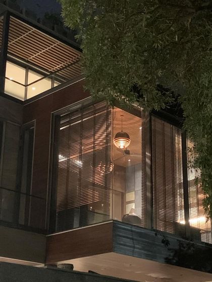 The cantilevered study seen from the outside at night, glowing like a lantern in the trees. The warm interior light highlights the intricate details of the pendant lamp and the rich wooden textures.