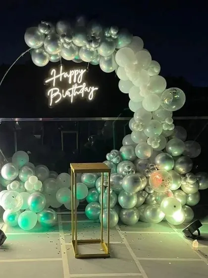 A simple and elegant balloon arch on a terrace. The silver and white balloons are arranged on a ring frame with a neon sign, creating a chic party backdrop.