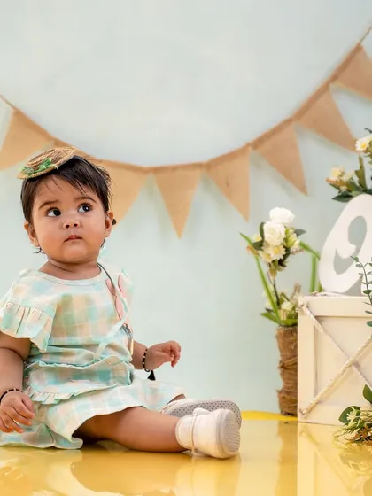 This little one, dressed in a sweet plaid dress, sits thoughtfully amidst her first birthday decorations in the studio.