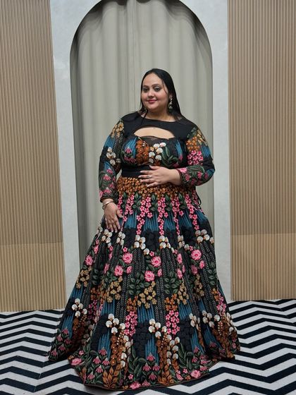 A plus-size model in a unique black lehenga with colorful floral embroidery. The keyhole neckline and full sleeves offer a modern yet modest look, available for rent.