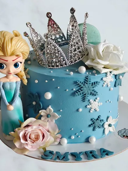 A chic and modern Frozen-themed cake for a 5th birthday. This design features a stylish Elsa figurine, a sparkling crown, and delicate sugar flowers, combining a popular theme with an elegant aesthetic.