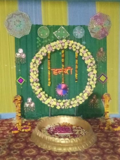 A traditional Haldi setup with a large urli and a backdrop of marigold strings. The decor uses a mix of yellow and light blue drapes with colorful hangings.