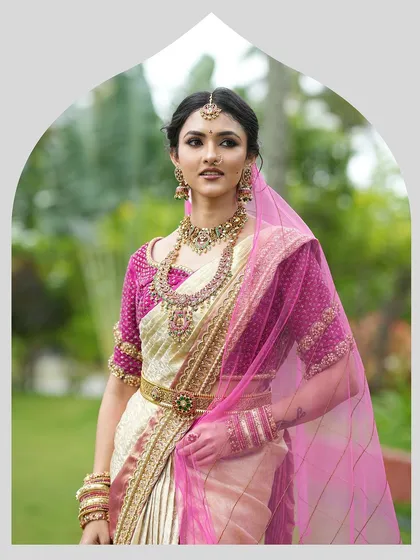 A classic South Indian bridal portrait. The bride is adorned in a cream and gold silk saree with a vibrant magenta blouse, complete with traditional jewelry and a sheer pink veil.