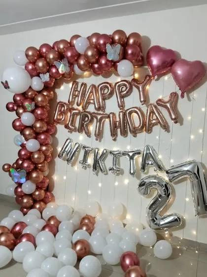 A pretty rose gold and white decoration for a 27th birthday. This setup includes a balloon arch, 'Happy Birthday' foil letters, and a wall of fairy lights.