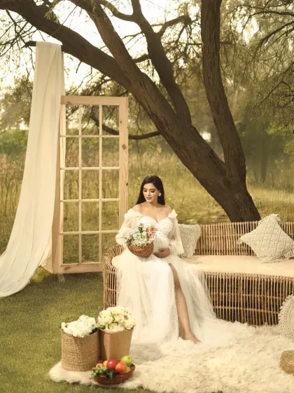 A serene moment from a boho-styled shoot. The mother-to-be sits on a wicker sofa, holding a bouquet, her expression peaceful and content.