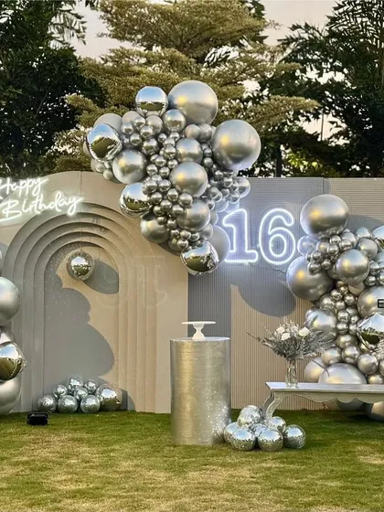A sleek and modern "Sweet Sixteen" birthday setup. The design features a minimalist grey backdrop with an arched detail, a neon "Happy Birthday" sign, and dramatic chrome silver balloon garlands.