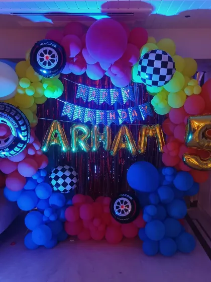 A Hot Wheels themed room decoration under party lights. The vibrant colors of the balloons and the shiny foil letters create an exciting atmosphere for a 5th birthday.