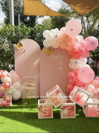 A delicate butterfly-themed birthday decoration in soft pink tones. This outdoor setup includes custom arched backdrops, balloon-filled transparent boxes spelling out the name, and a balloon-filled marquee number.