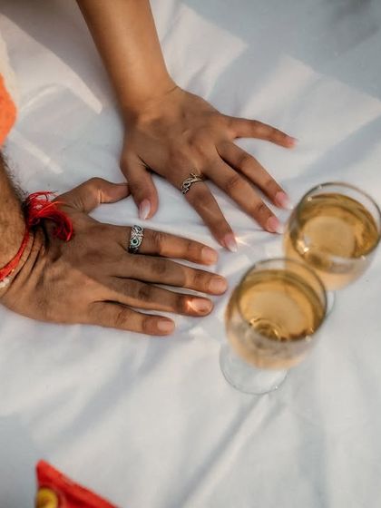 A detail shot of the couple's hands, adorned with their rings, resting on the picnic blanket next to two glasses of wine, symbolizing their shared life and celebration.