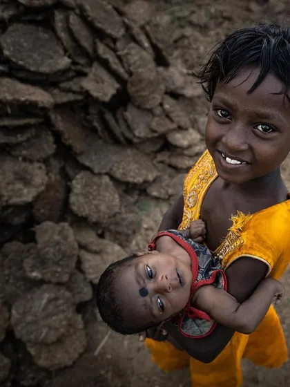 In a rural village, an older sister holds her sibling, her bright smile a stark contrast to the earthy tones of her surroundings. This is a simple, honest portrait of human connection, joy, and the responsibilities of childhood.