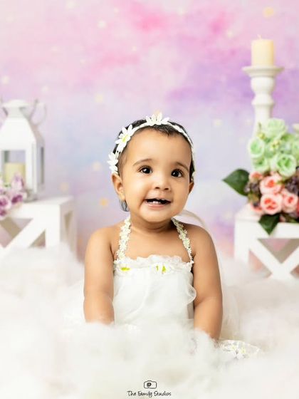 A happy baby girl surrounded by soft clouds and flowers in a whimsical studio setup.