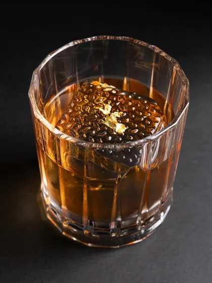 A close-up of a whiskey-based cocktail with a unique, textured ice cube and a fleck of gold leaf. The dark, moody lighting gives it an ultra-premium and sophisticated look.