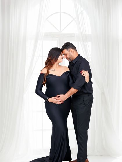 An intimate moment between an expecting couple, captured in a clean and bright studio setting. The focus is on their connection as they embrace before the bright window.