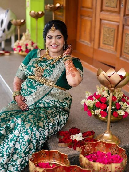 A happy mom-to-be seated amidst flowers at her baby shower. Her smile and the beautiful henna on her arms capture the festive and joyous spirit of the occasion.