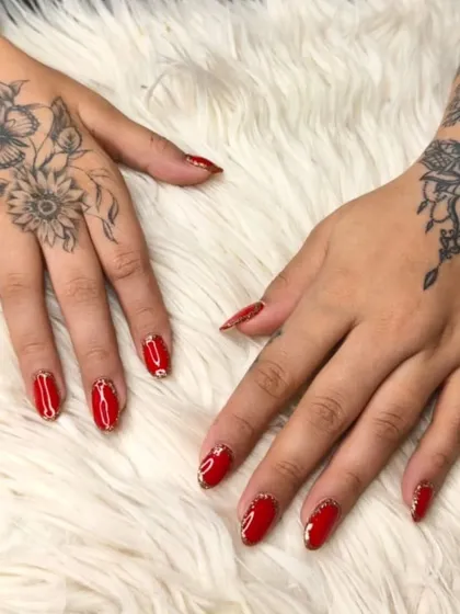 Vibrant red gel polish on oval nails, enhanced with a gold glitter cuticle accent and delicate stone art for a touch of luxury.