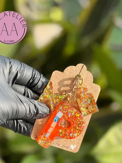 A close-up of a custom initial keychain. This one, for a friend named Rita, features a vibrant orange color with gold glitter and a matching charm.