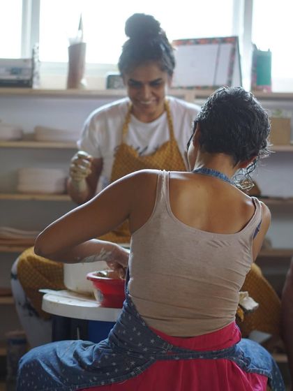 A beautiful shot of two students working at their wheels, with one smiling in the background, capturing the supportive and happy atmosphere of the studio.