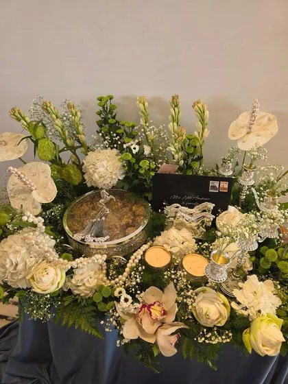 An opulent arrangement in white and silver. This hamper features a silver dry fruit box, pearl strings, and decorative silver pieces surrounded by white roses, anthuriums, and tuberoses.