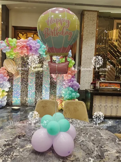 A hot air balloon themed table centerpiece. A large foil balloon floats above a small cluster of pastel balloons, tying the overall theme together across the venue.
