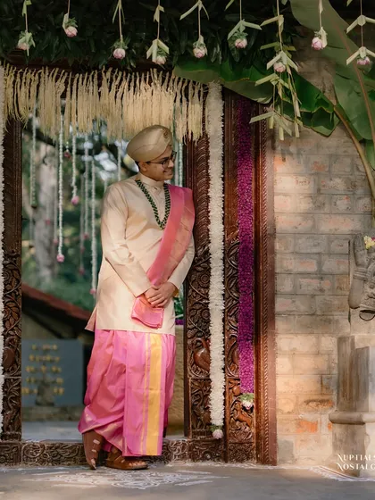 The groom, in his traditional pink and cream wedding attire, waits by a decorated doorway, a picture of anticipation.