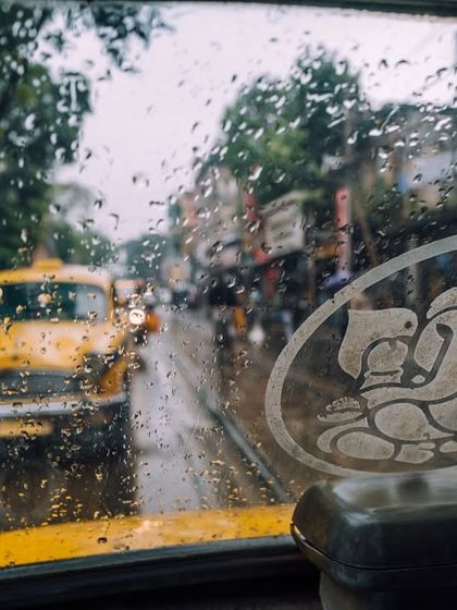The view from inside a classic yellow Ambassador taxi on a rainy day in Kolkata. The raindrops on the windshield and the Ganesha sticker create a uniquely Kolkata mood.