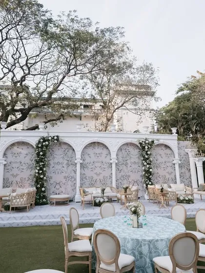 For this outdoor event, I designed a sophisticated backdrop with classic white arches and a delicate floral wallpaper pattern, complemented by elegant lounge furniture.