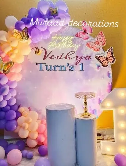 This butterfly-themed first birthday is soft and dreamy. The setup includes a pastel rainbow balloon garland, a large light-up number '1', and delicate butterfly accents on a circular backdrop.