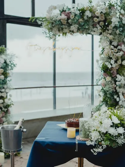 A closer look at the proposal setup, featuring a table for two with cake and champagne, all set against the stunning backdrop of the sea and a lush floral arch.