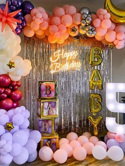 A "Baby" shower or first birthday setup with a silver fringe backdrop. The decoration features 'BABY' blocks, a marquee number '1', and a multi-colored balloon garland.