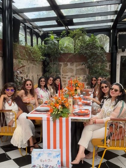 The ladies enjoying a beautiful brunch at the Hermès-themed baby shower. The long table is decorated with vibrant orange floral runners and personalized place settings.