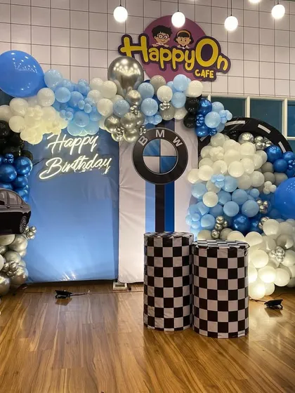 A fun and dynamic BMW car-themed birthday decoration for a 5th birthday. The setup includes a balloon arch in blue, black, and silver, checkered racing flag pedestals, a car cutout, and the iconic BMW logo for a young car enthusiast.