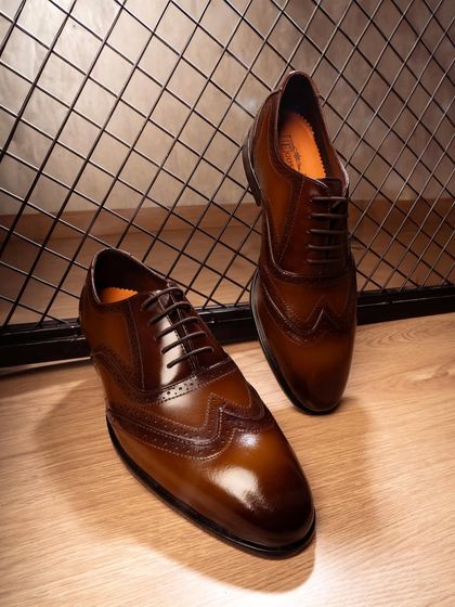 A pair of classic brown brogues against an industrial mesh background. The warm lighting and setting give these formal shoes a modern, stylish edge.