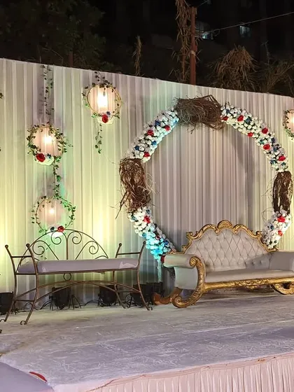 This elegant stage decor features a central floral ring, a classic white and gold sofa, and hanging floral lanterns against a draped white backdrop. The design is perfect for a sophisticated engagement or reception.
