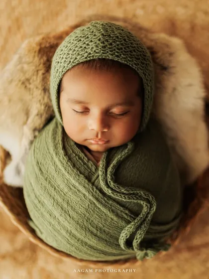 Earthy green tones look so beautiful on newborns. This baby is peacefully asleep in a basket, wearing a soft green wrap and bonnet that complements the natural textures of the setup.