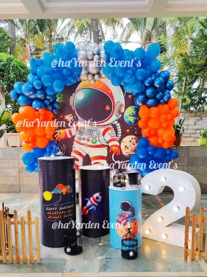 An outdoor space-themed party for a second birthday. The astronaut backdrop is surrounded by a vibrant blue and orange balloon arch.