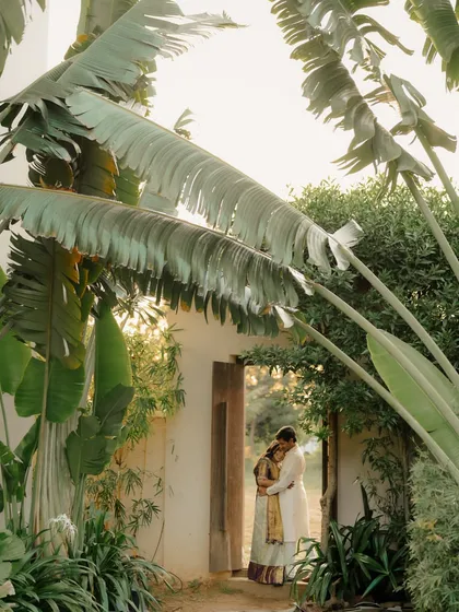 A beautifully framed shot of a couple embracing in a doorway, surrounded by large, tropical leaves. The natural frame creates a sense of intimacy and seclusion.