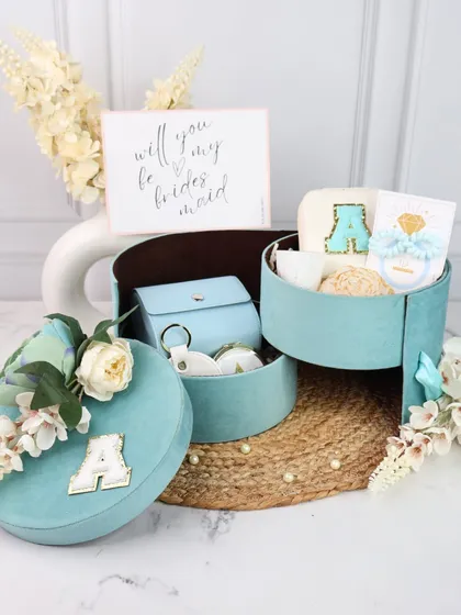 A "Will you be my bridesmaid?" proposal hamper in a stunning turquoise suede box. This round, tiered box is personalized with an initial and filled with custom accessories for your bridal crew.