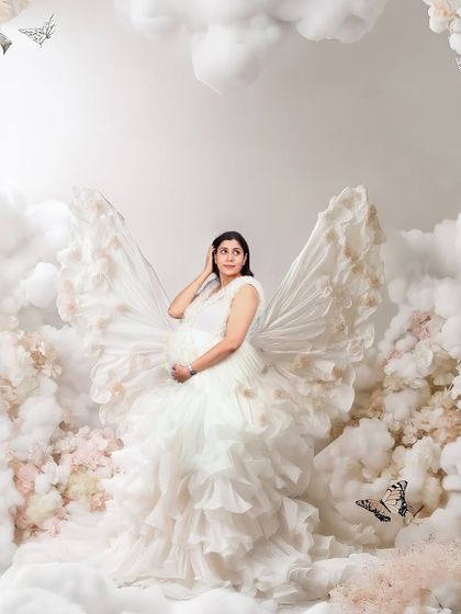 An ethereal portrait of a mother-to-be with butterfly-like wings, standing amidst a sea of clouds and flowers. This heavily edited, dreamy image creates a magical, fairy-tale scene.