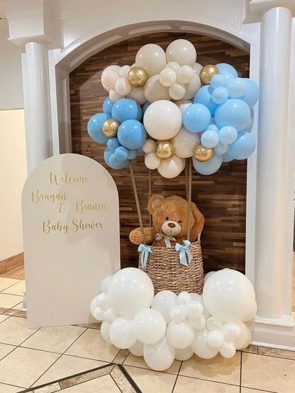 A charming baby shower setup with a hot air balloon basket prop holding a teddy bear, surrounded by blue and white "cloud" balloons.