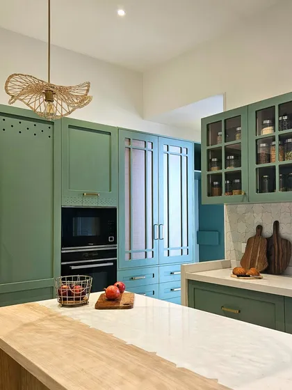 A wider view of the soft green kitchen, highlighting the unique island that combines wood and marble. The artisanal pendant light and glass-fronted cabinets for spices add to the room's layered and thoughtfully curated feel.