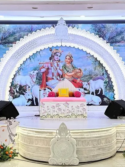 The main stage for a seven-day Bhagwat Sapta event at Hiravati Hall. We created a serene all-white setup with an ornate arch framing a Radha-Krishna backdrop, complemented by decorative sun-like motifs and Pichwai cows.