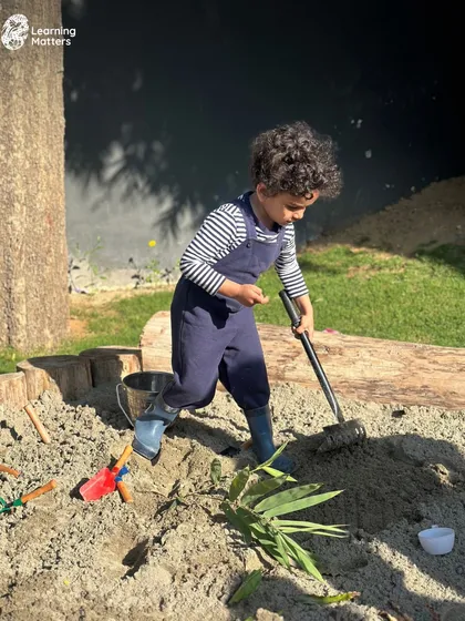 A child dressed as a gardener tends to the sandpit. Our outdoor spaces, including our sand and mud kitchens, are rich environments for imaginative play and connecting with natural elements.