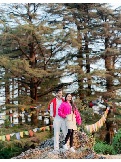 A vibrant, joyful shot from a pre-wedding session in the hills. The colorful prayer flags and towering pines create a beautiful, natural setting. This image captures the adventurous and free-spirited side of the couple's relationship.