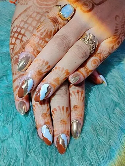 A unique design combining gold chrome, brown polish, and white abstract swirls, shown against a henna-adorned hand.